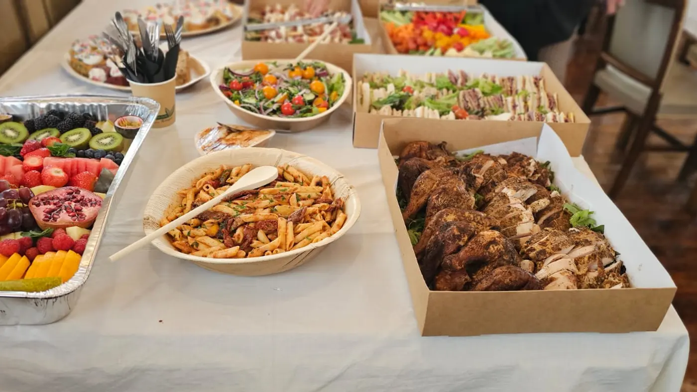 A buffet spread featuring a vibrant fruit platter, pasta salad, and carved chicken.