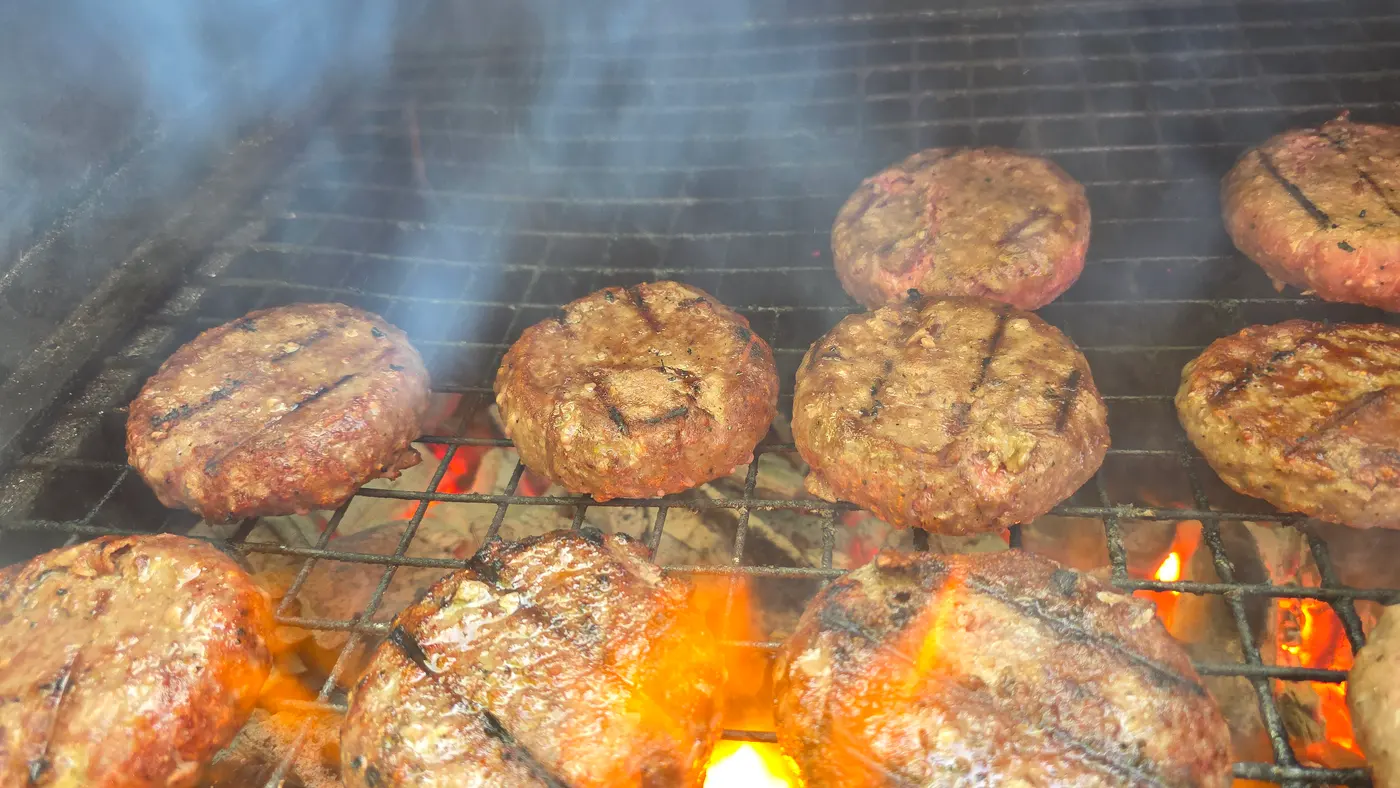 Juicy burgers are grilling over hot coals with visible smoke and flames.