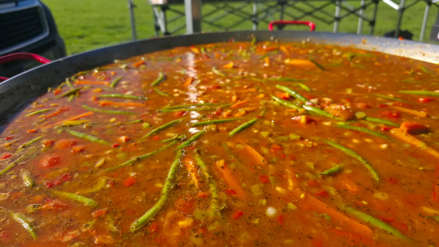 A large pan of paella cooking outdoors at an event with people in the background.