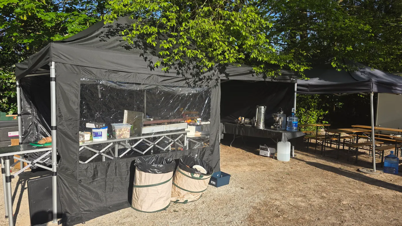Outdoor catering setup with tents, tables, and drink dispensers under trees.
