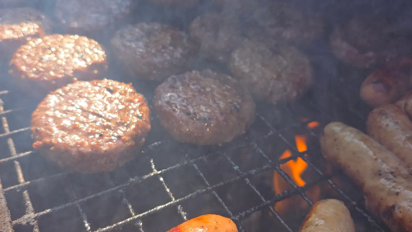 Burgers and sausages cooking on a smoky barbecue grill with visible flames.