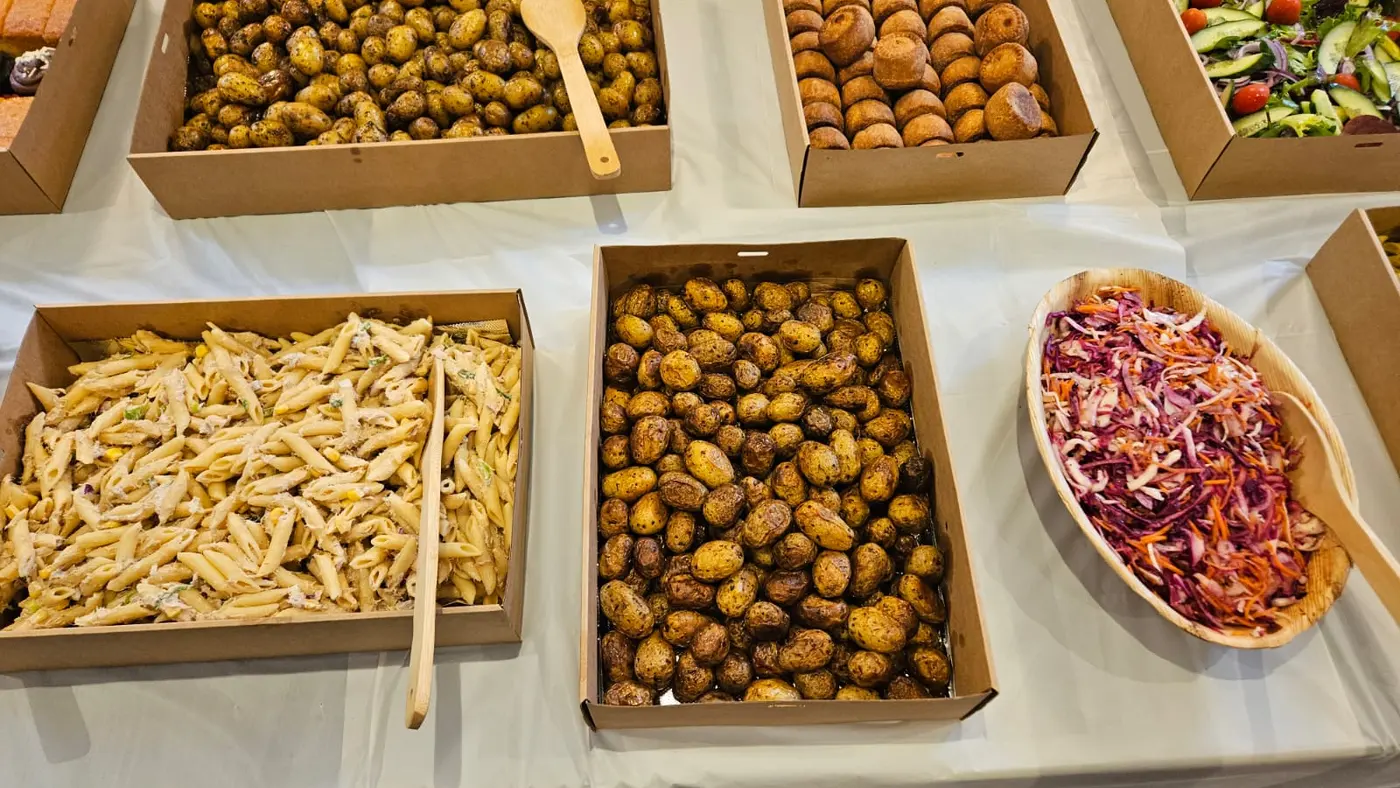 A buffet spread features trays of roasted potatoes, pasta salad, coleslaw, and Yorkshire puddings.