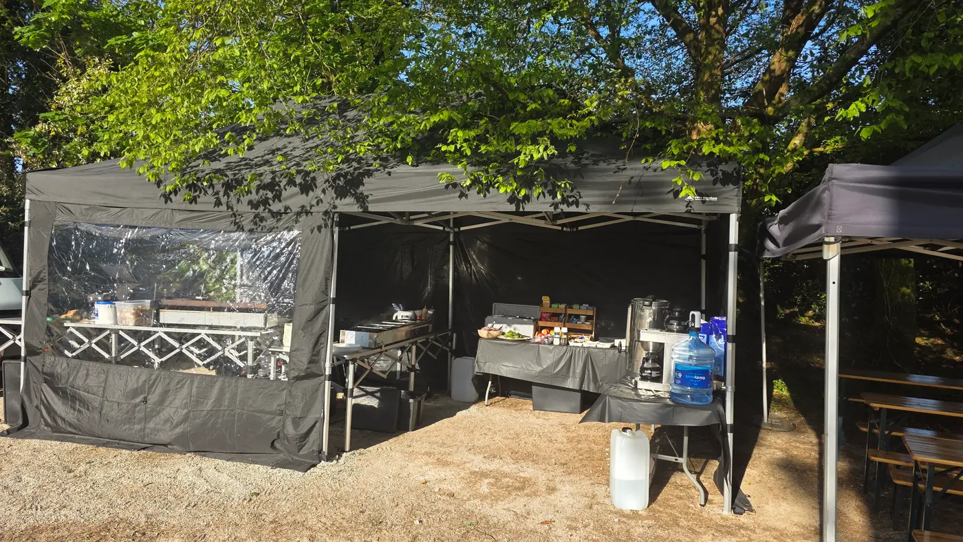 Outdoor catering setup with tents, tables, and food service equipment under trees.