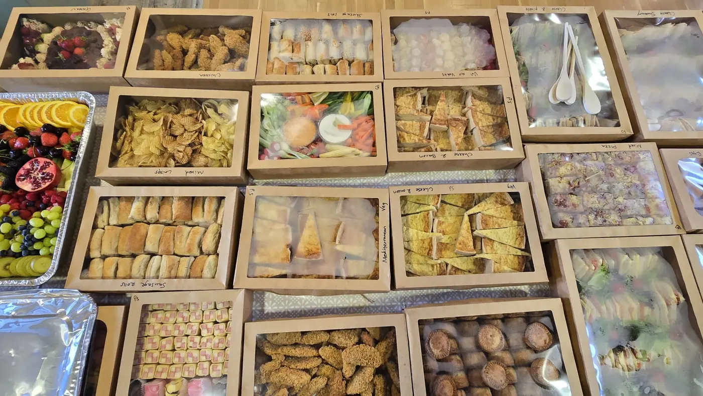A variety of catering boxes including fruit, sandwiches, pastries, and quiches.