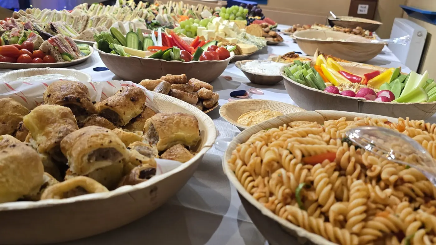 A long table laden with a diverse buffet of sandwiches, sausage rolls, pasta, and crudités.