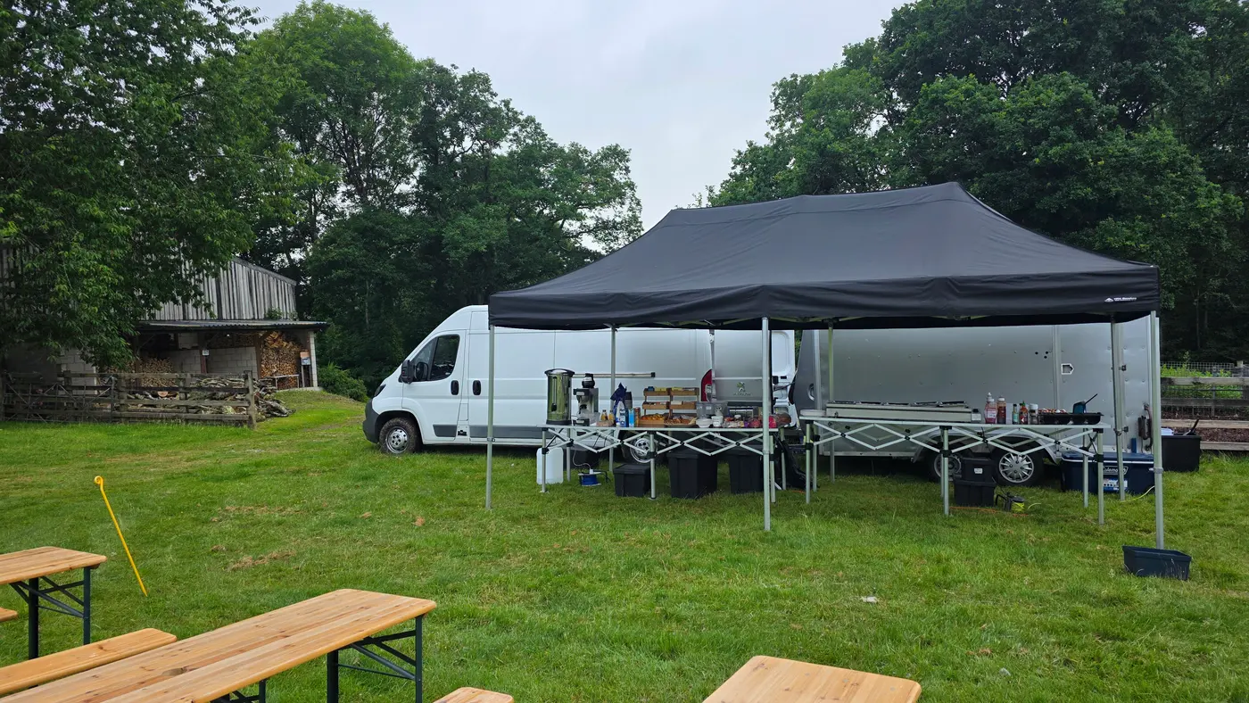 Outdoor catering setup with van, trailer, gazebos, and picnic tables.