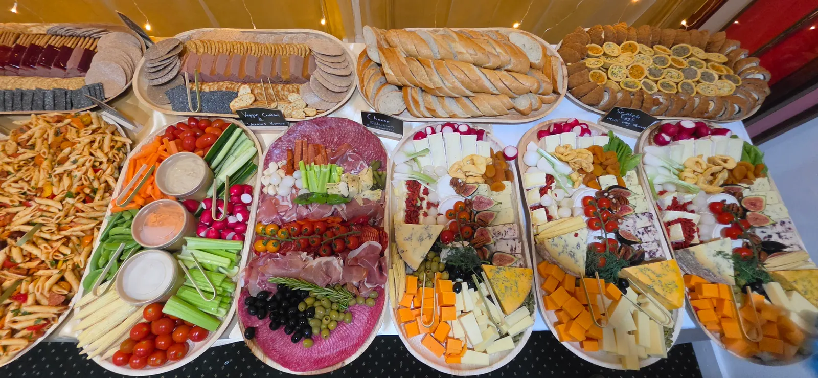 Cured meat board with prosciutto, salami and chorizo beside four cheese boards, vegetable crudites with dips, scotch eggs, mini quiches and sliced baguettes.