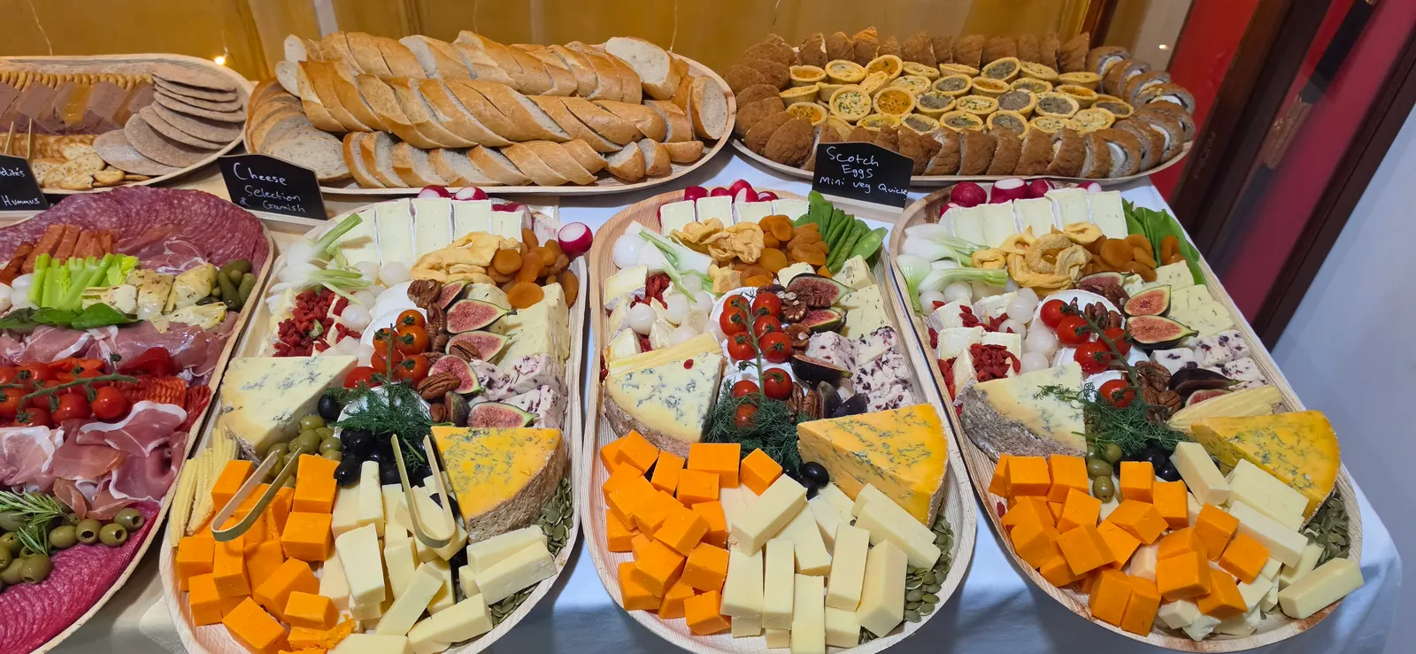 Three matching cheese boards side by side, each with blue cheese, cheddar, brie, figs, dried apricots and pecans, behind trays of sliced baguette, scotch eggs and mini vegetable quiches.
