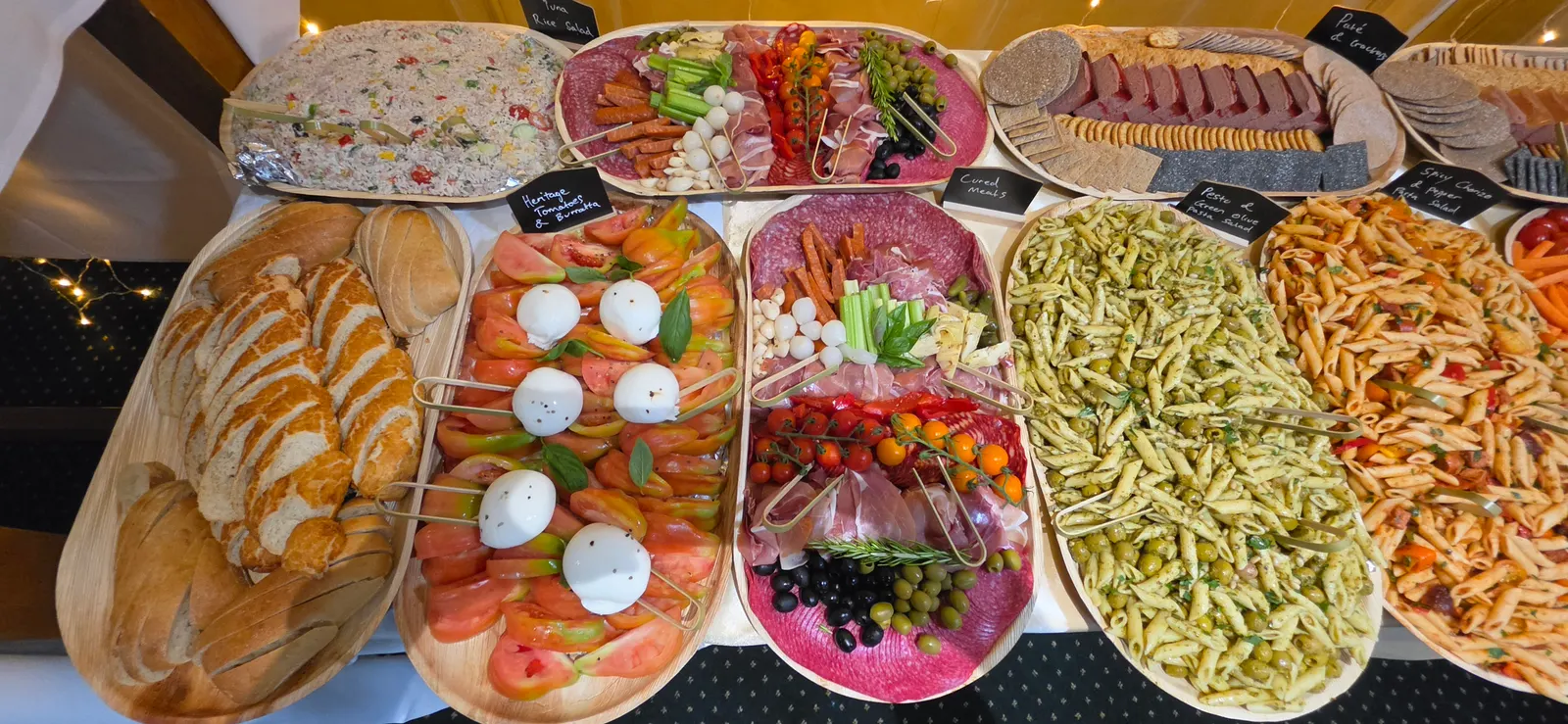 A board of heritage tomatoes and burrata with basil, beside sliced breads, cured meats, tuna rice salad and two pasta salads, each board marked with a chalkboard label.