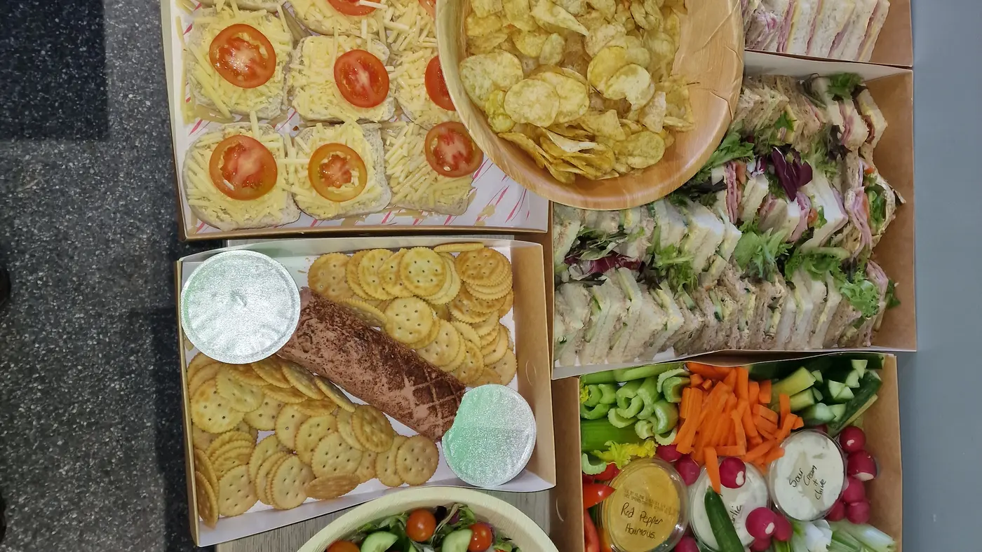 A selection of catering platters including sandwiches, salad, sausage rolls, and crudités.