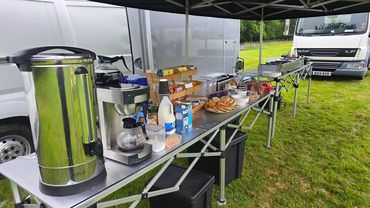 Outdoor catering setup with coffee, pastries, and drinks under a tent.