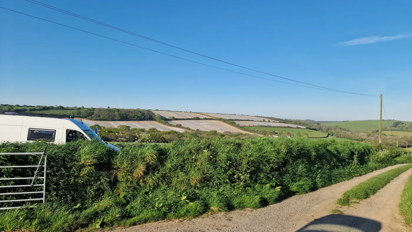 A white van parked beside a hedgerow in a rural Cornish landscape.