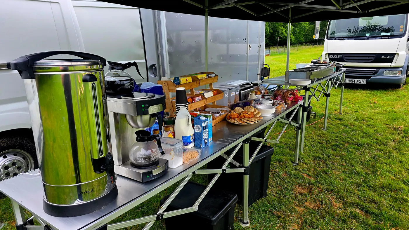 Outdoor catering setup with coffee, pastries, and buffet items under a tent.