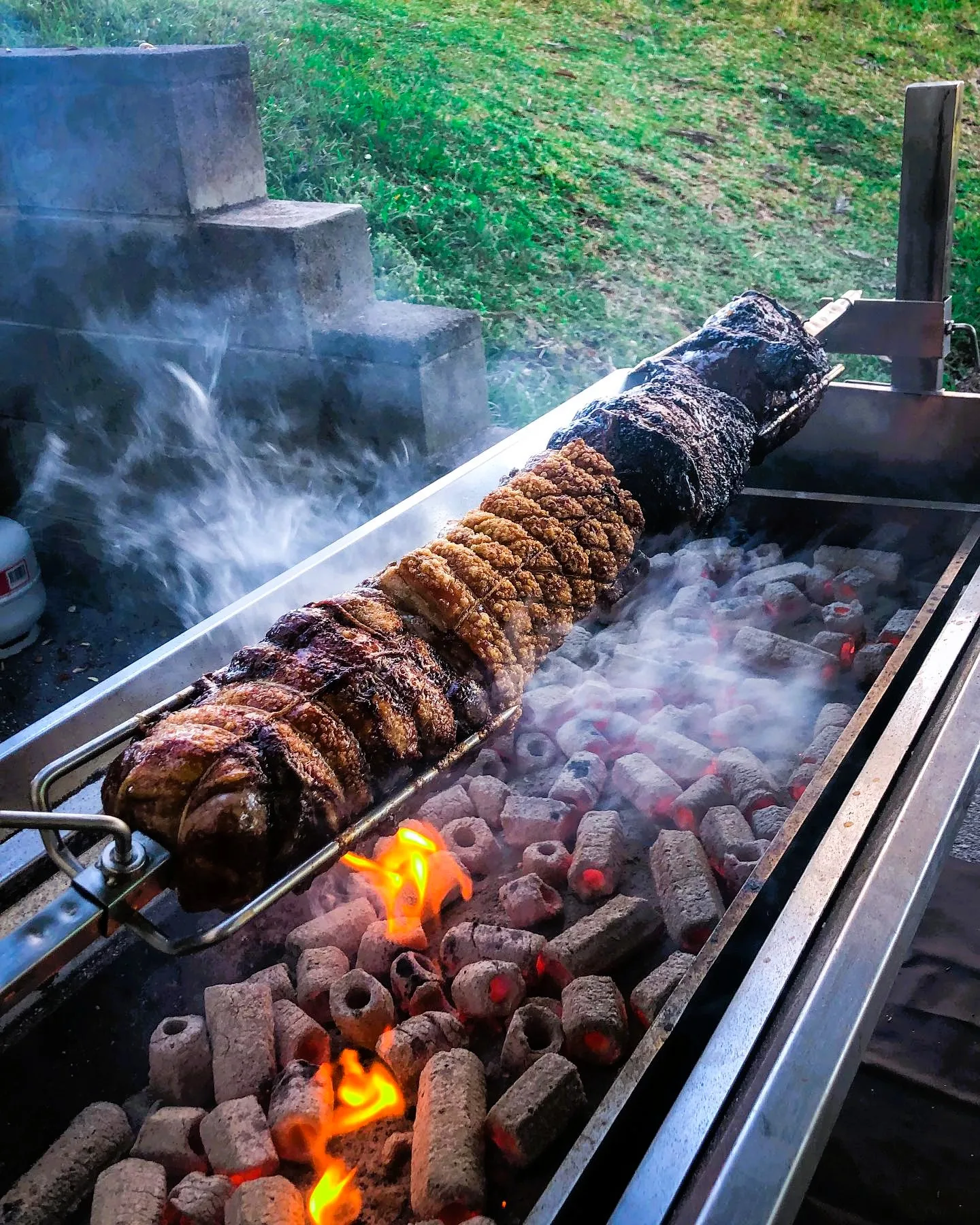 Hog turning slowly over glowing briquettes with rising smoke at an outdoor event