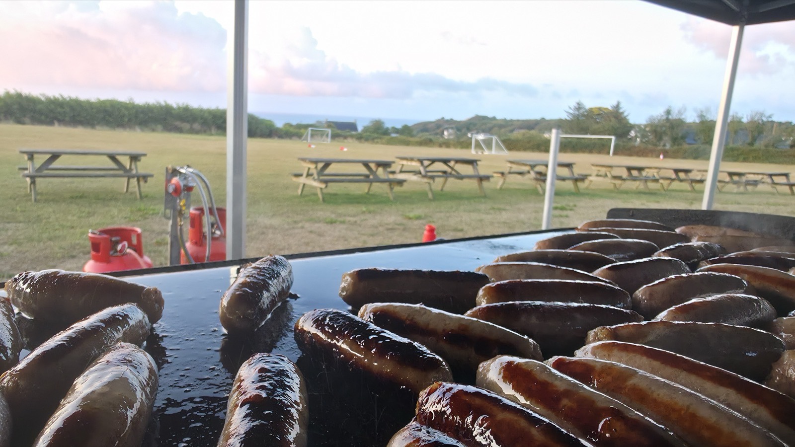 Breakfast cooking on a griddle under a gazebo in a field above Cadgwith at dawn, gas bottles alongside